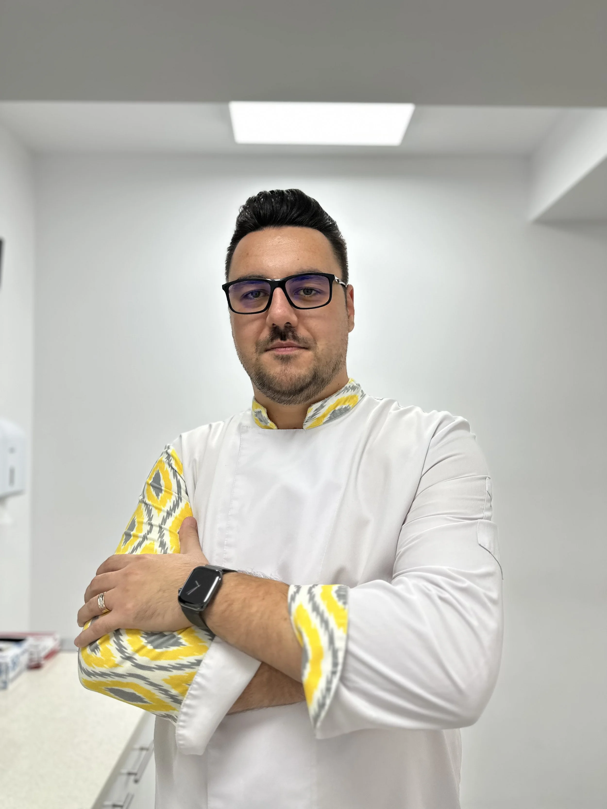 Man wearing glasses and a white lab coat with a colorful patterned shirt underneath, standing with arms crossed in a clinical setting.