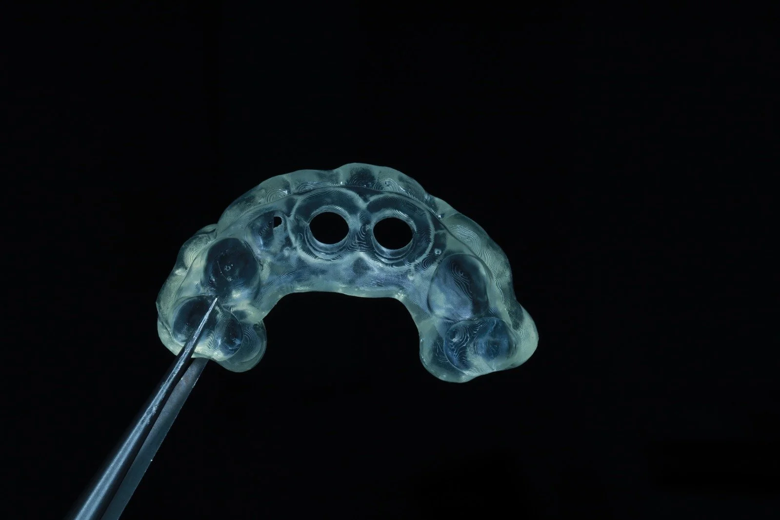 A transparent dental implant surgical guide held by tweezers against a black background.