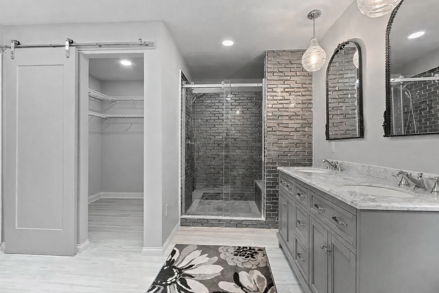 A modern bathroom with a double vanity with marble countertop, two mirrors, pendant lights, a glass shower with brick tiles, and an open closet with wire shelving.