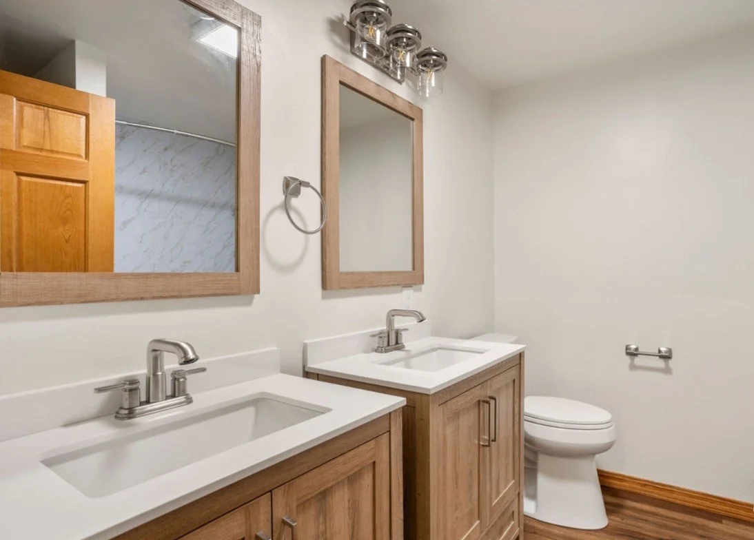 Bathroom with double vanity, two mirrors, two sinks, towel ring, toilet paper holder, and toilet, with wood accents and a wood floor.