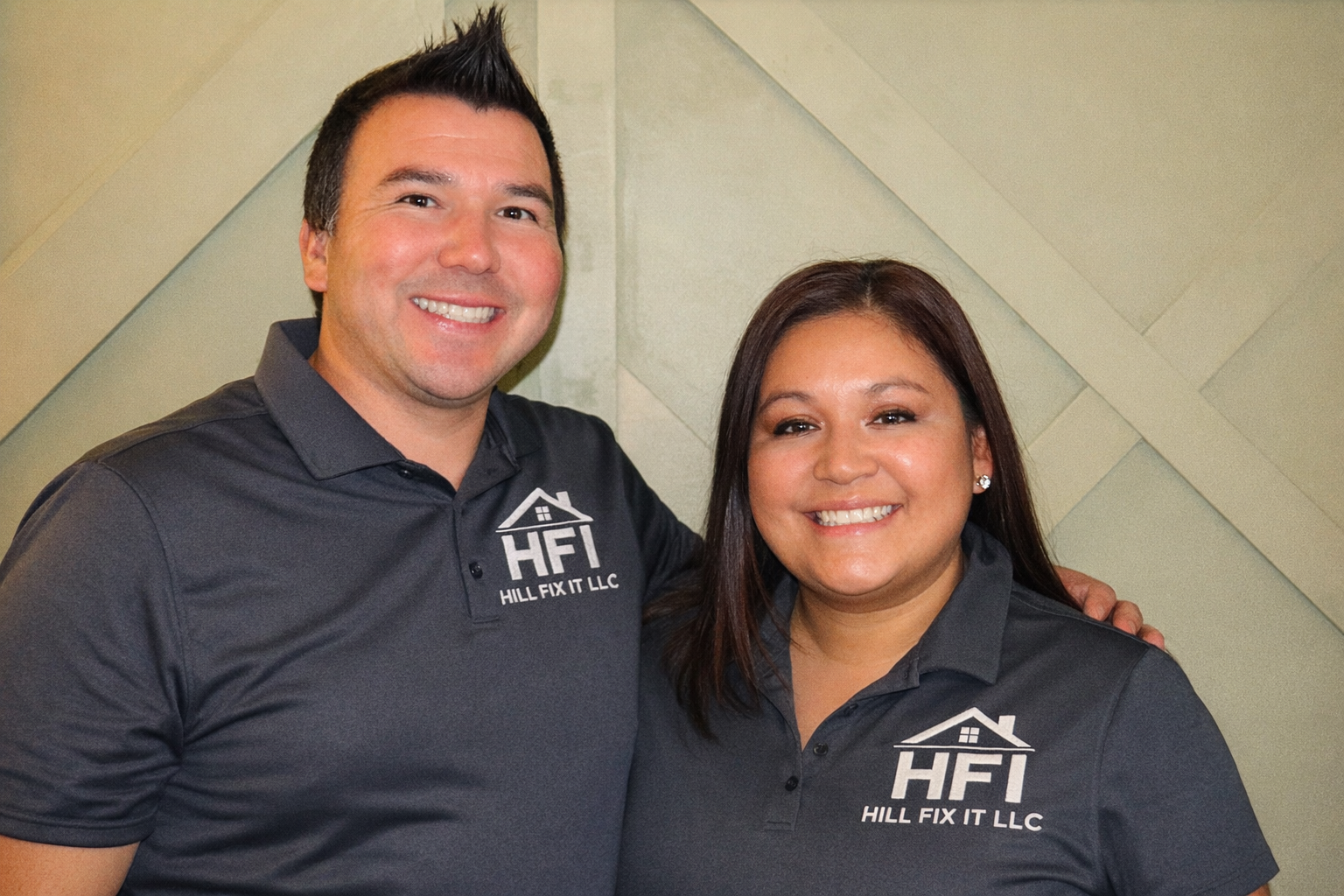Two smiling individuals wearing matching dark grey polo shirts with a house logo and the text "HFI Hill Fix It LLC." They are standing against a light-colored textured wall, with the person on the left having short dark hair styled upward and the person on the right having shoulder-length dark hair.