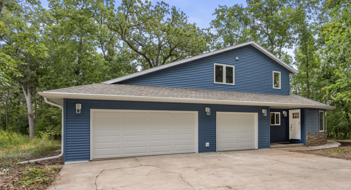 A two-story blue house with a white garage door, situated among trees, with a concrete driveway and a small front porch.