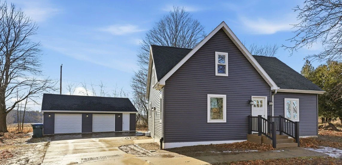 A two-story house with dark purple siding, white trim, and a small front porch with stairs, alongside a detached three-car garage on a clear day with some snow on the ground.