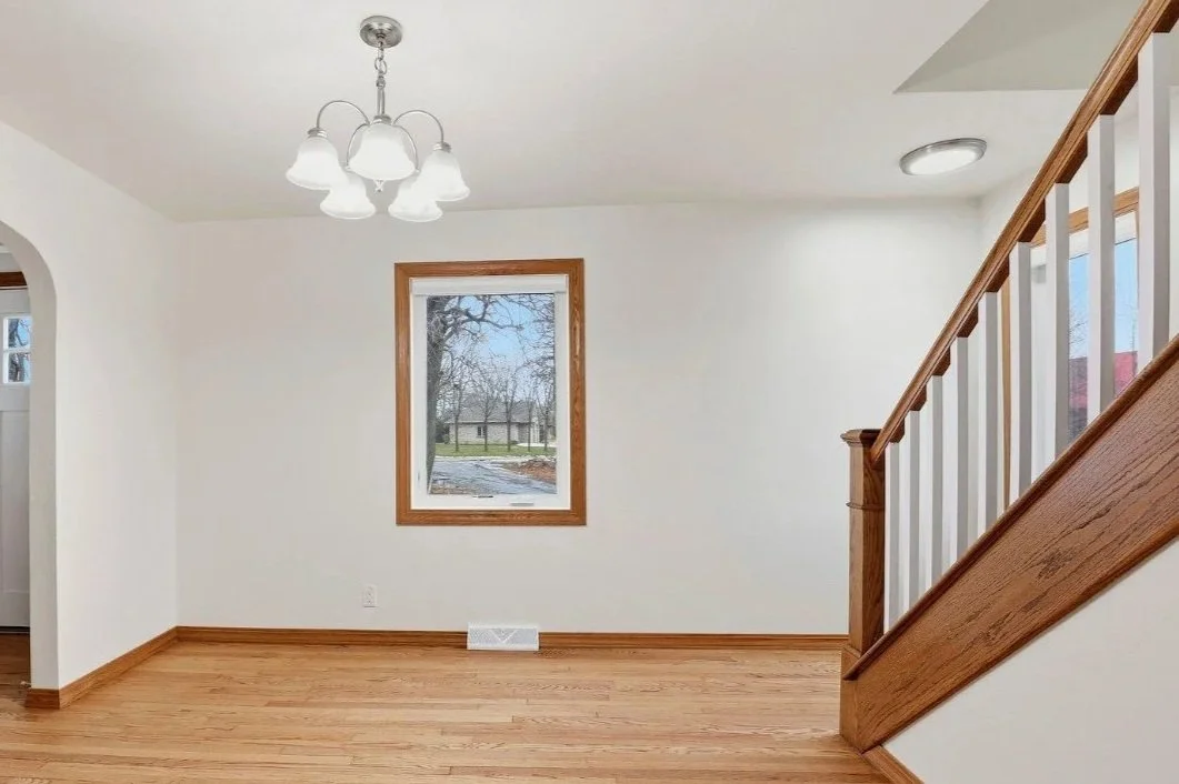Empty room with white walls, wooden baseboards, a window with a view of trees, a wooden staircase with white balusters, and a chandelier with five frosted glass shades.