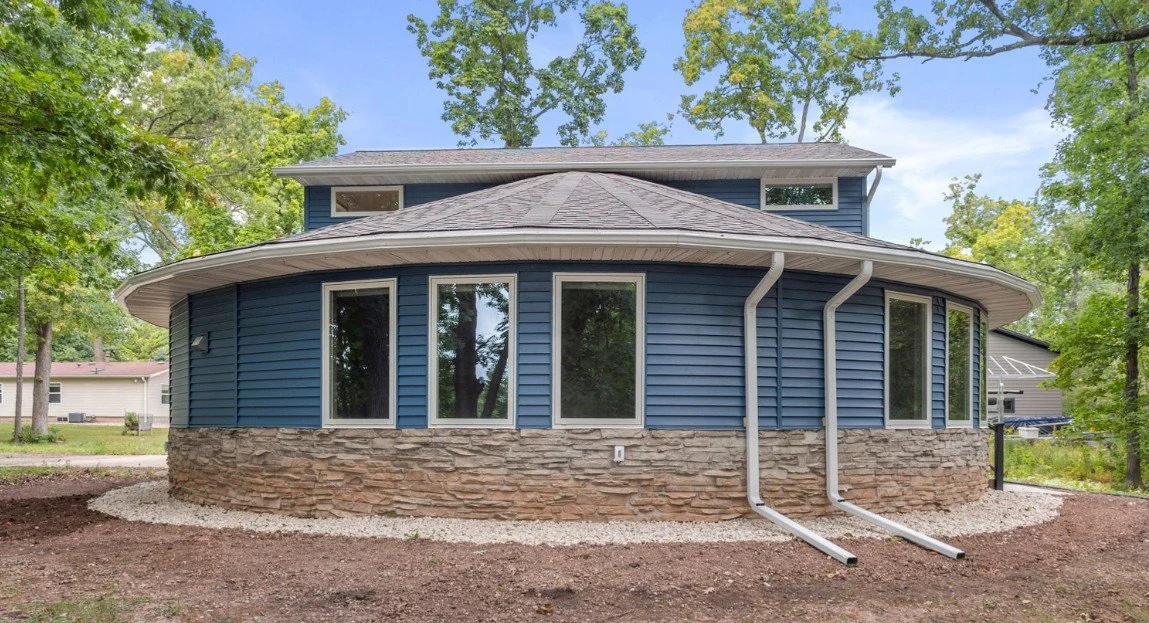 Blue house with curved wall, stone foundation, multiple large windows, and surrounding trees.