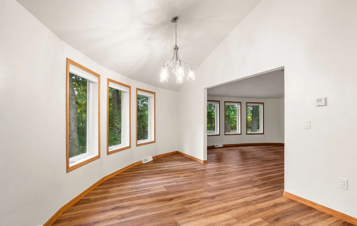 Empty living room with large windows overlooking trees, hardwood floors, and a modern chandelier.