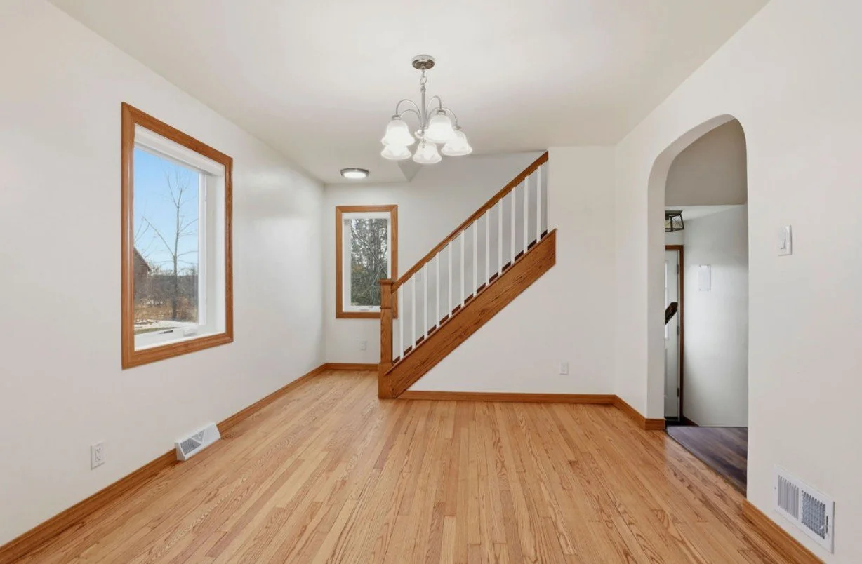 Empty living room with hardwood floors, white walls, two windows with wooden trim, a staircase with wooden railing, a chandelier, and an arch entryway.