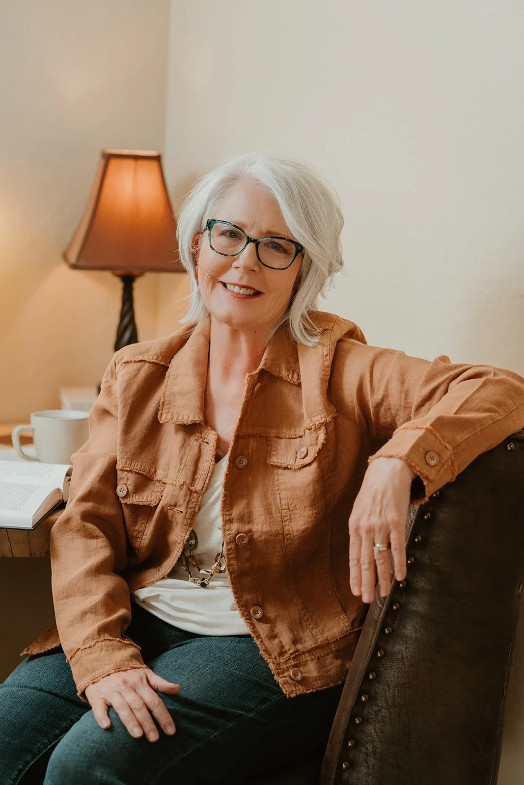 An older woman with glasses and gray hair smiling while sitting on a dark brown leather chair in a cozy room, with a lamp, mug, and open book on the table behind her.