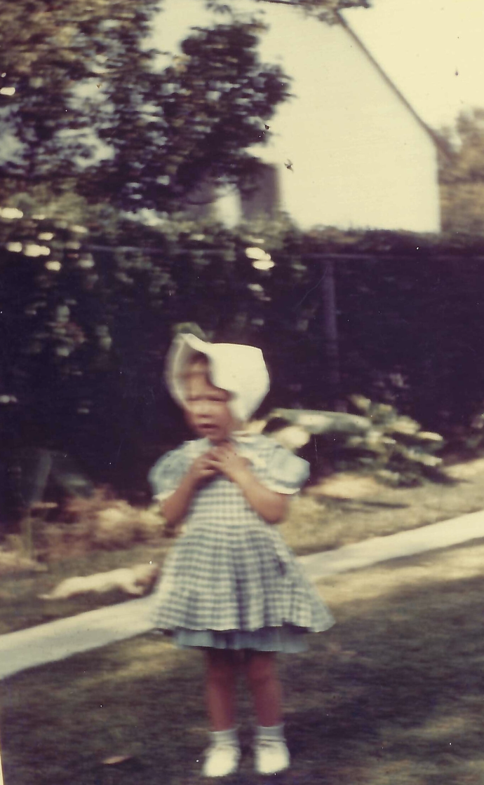 Young girl wearing a checkered dress and headscarf standing outdoors on a lawn.