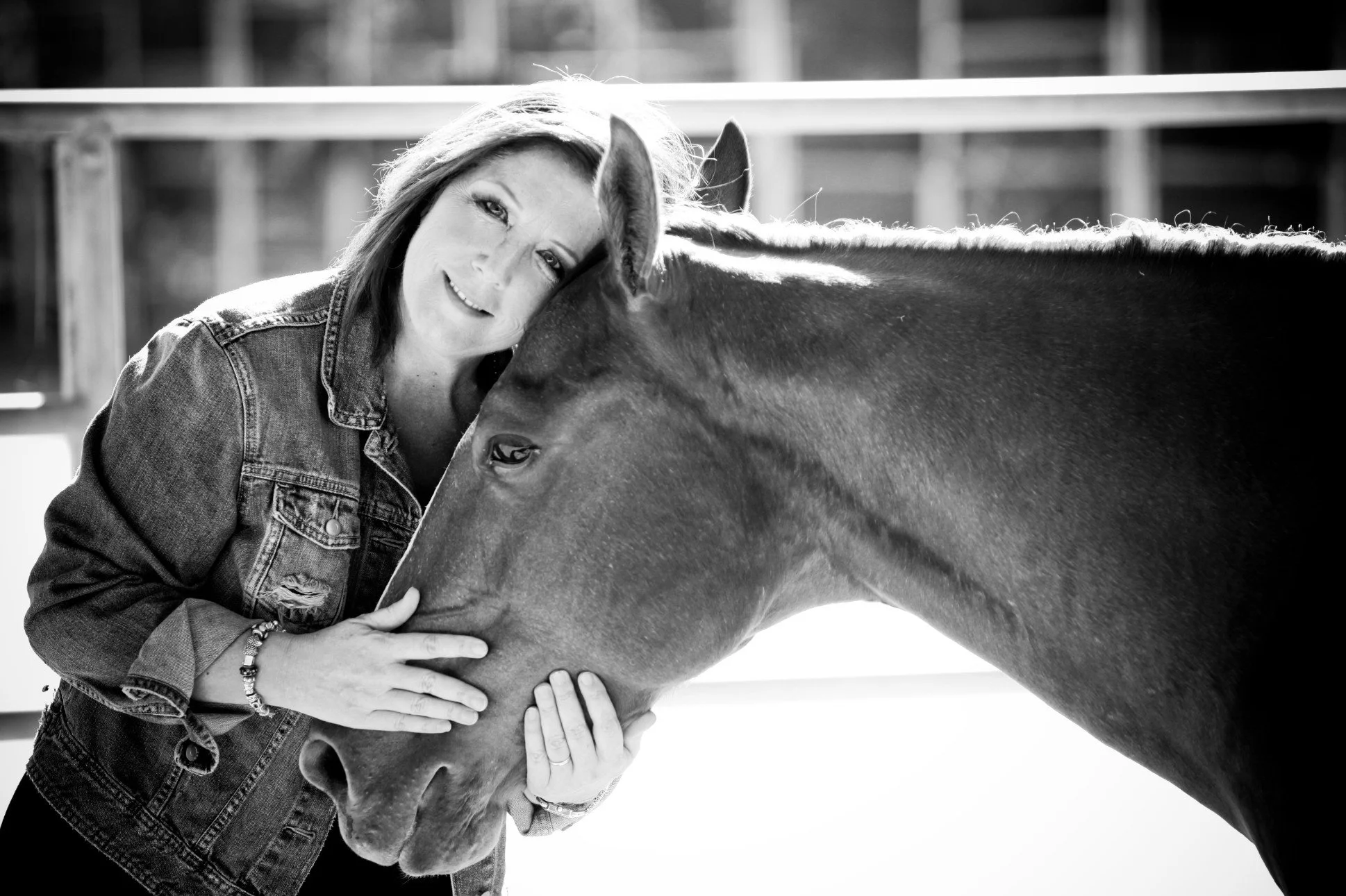 A woman with shoulder-length hair, wearing a denim jacket, is gently resting her head and hand on the head of a horse, both appearing affectionate. The woman is smiling softly, and the horse's eyes are calm. The background shows part of a wooden fence, indicating an outdoor setting.