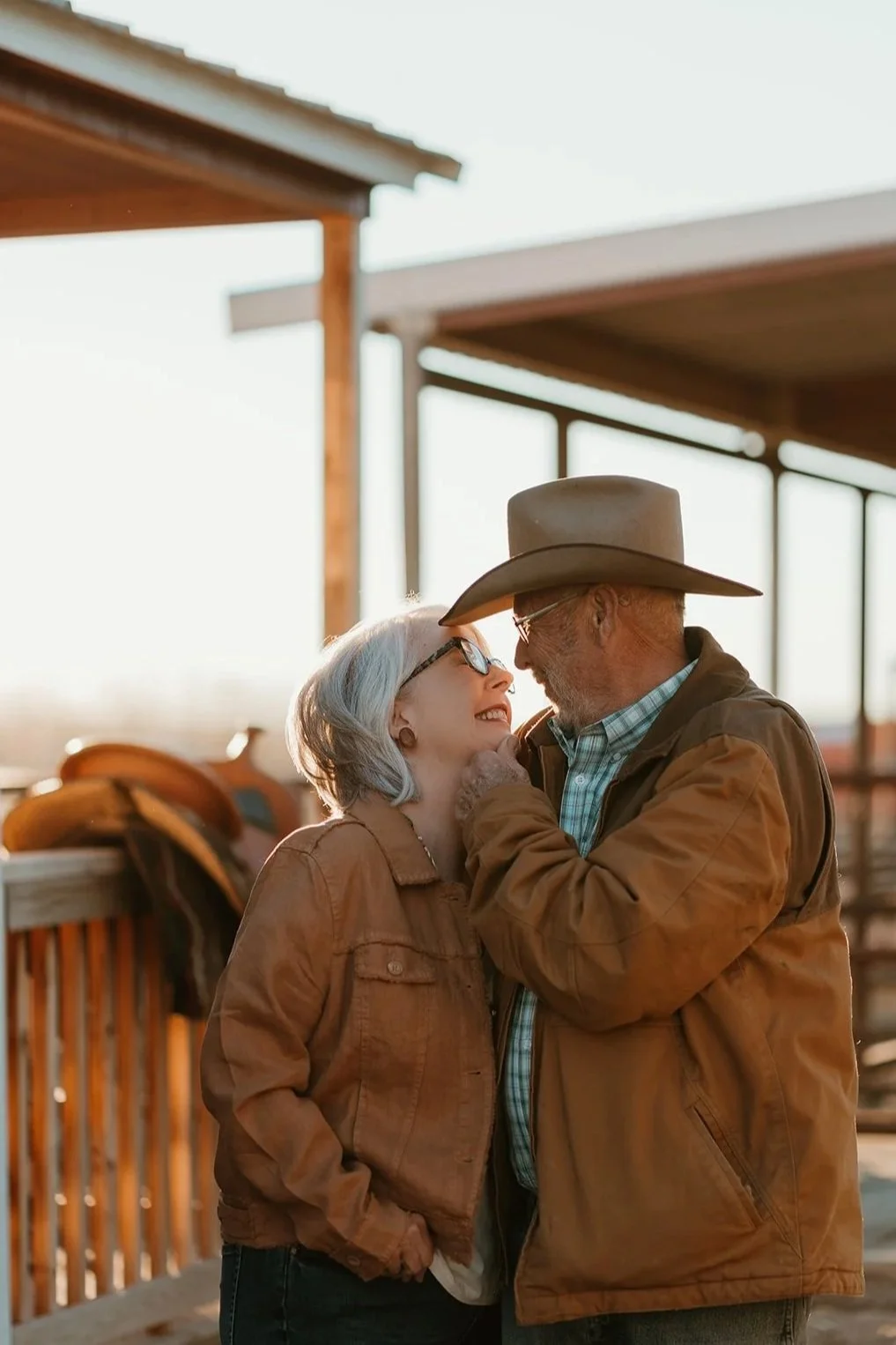 An elderly couple smiling and looking at each other lovingly, standing close together outdoors in a rustic setting with wooden structures and a saddle in the background, during sunset.