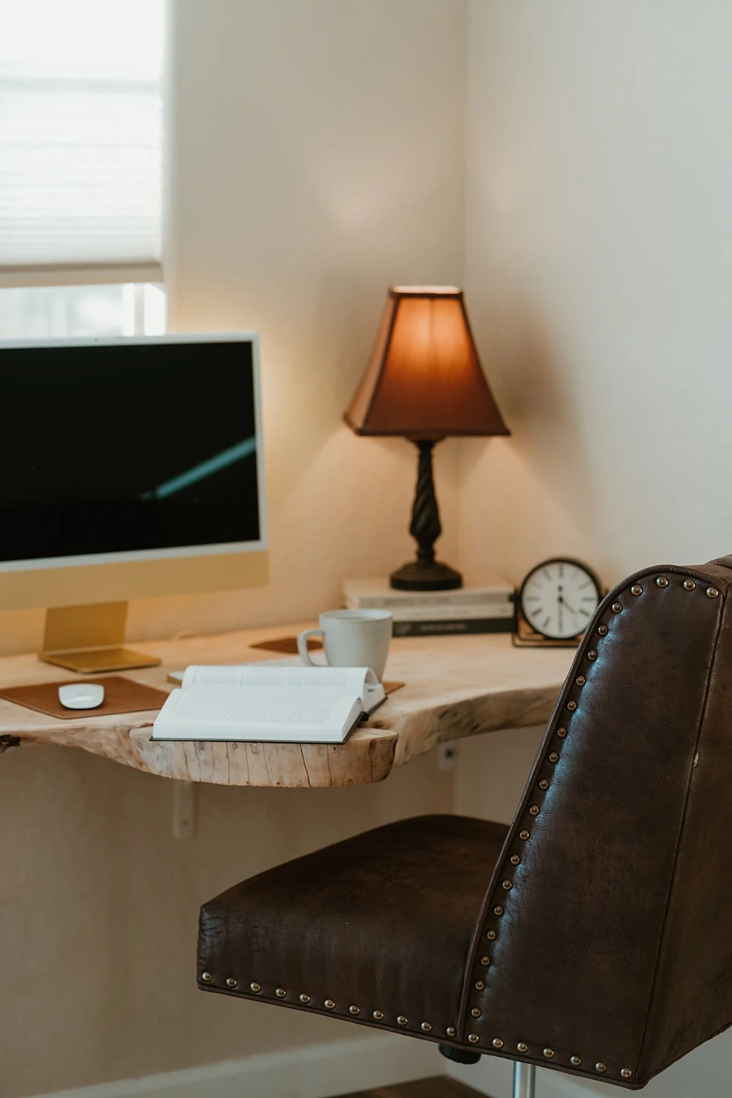 Home office desk with a computer monitor, open book, white coffee mug, desk lamp, clock, and stack of books; brown leather chair in foreground.