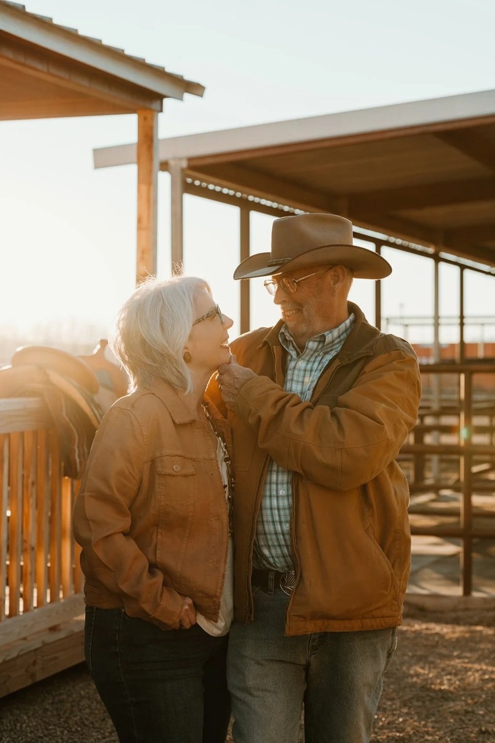 An elderly woman and man smiling at each other outdoors at sunset, with a wooden building and horse in the background.