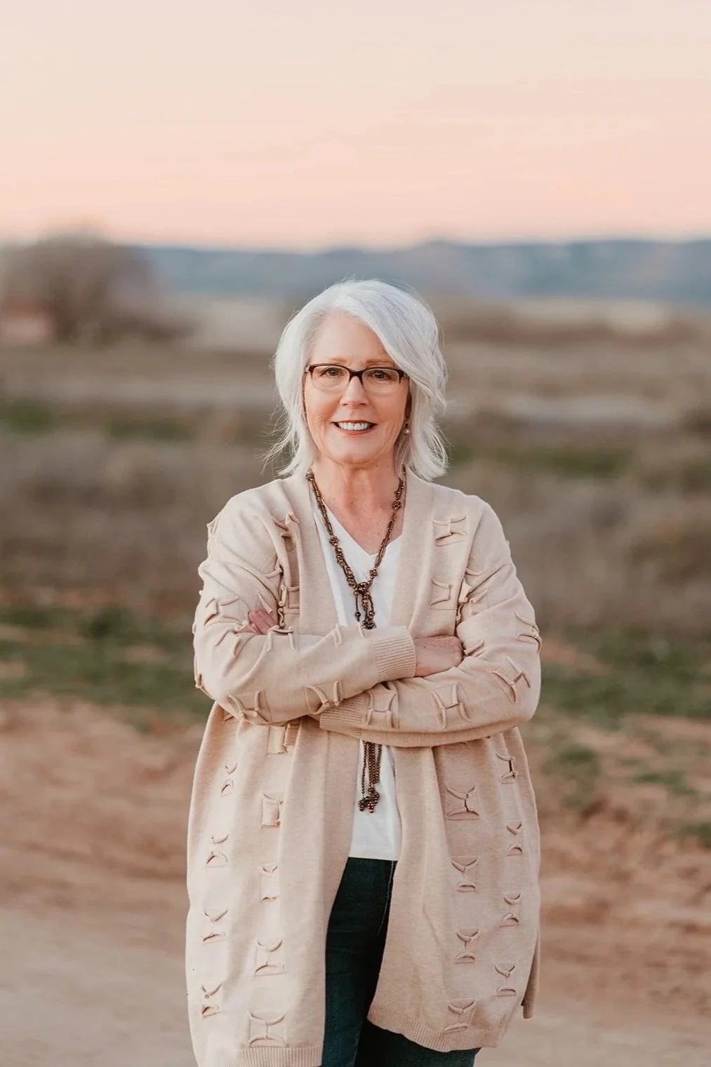 A smiling elderly woman with white hair and glasses, wearing a beige cardigan with cutouts and a necklace, standing outdoors with arms crossed against a blurred natural landscape at sunset.