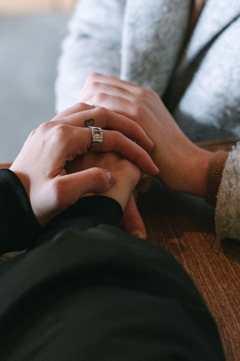 Two people holding hands on a wooden table, one person wearing a silver ring and a black jacket, the other wearing a beige sweater.