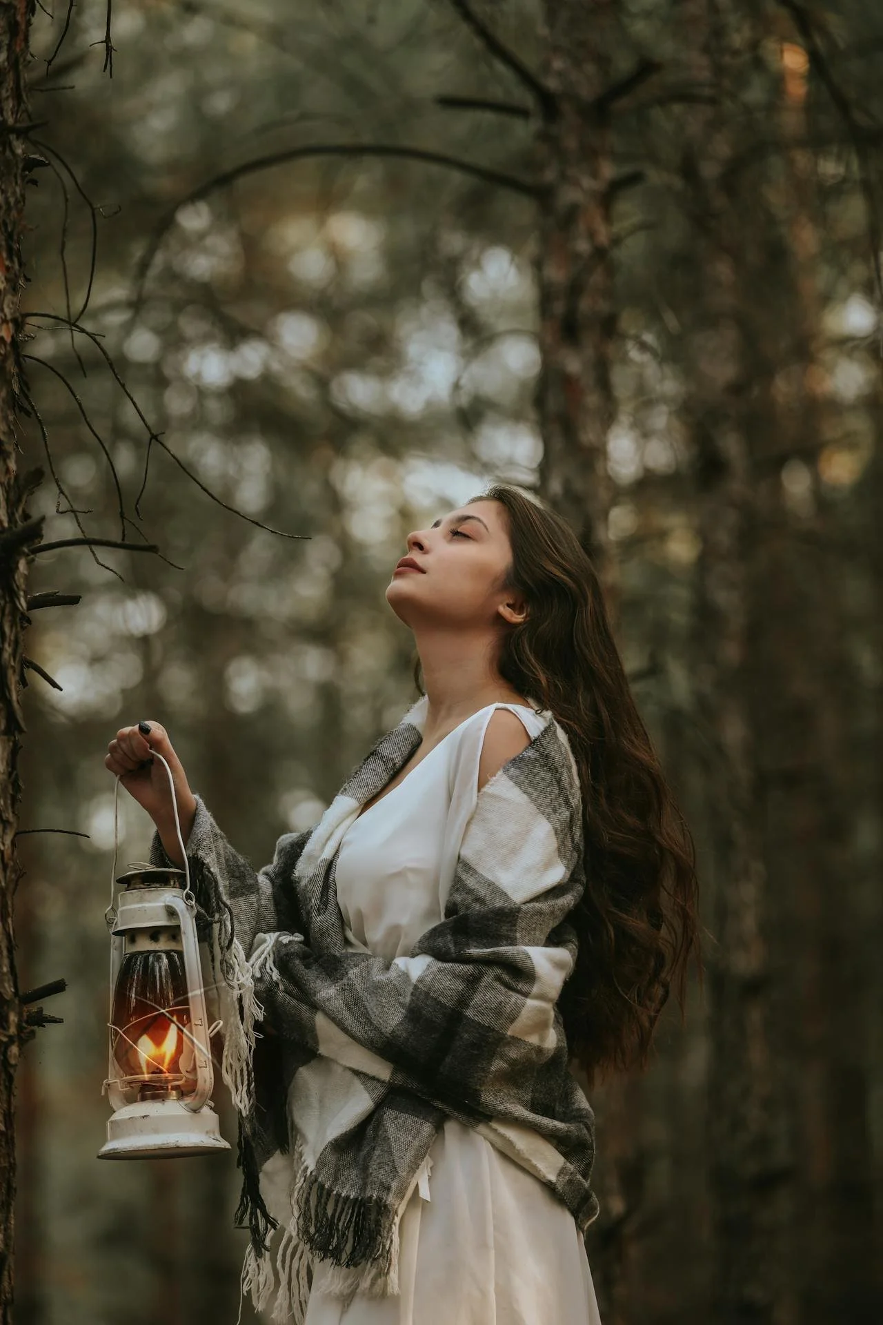 A woman with long, wavy brown hair holds a lantern and stands in a forest, with her eyes closed and head tilted back slightly, enjoying the moment.