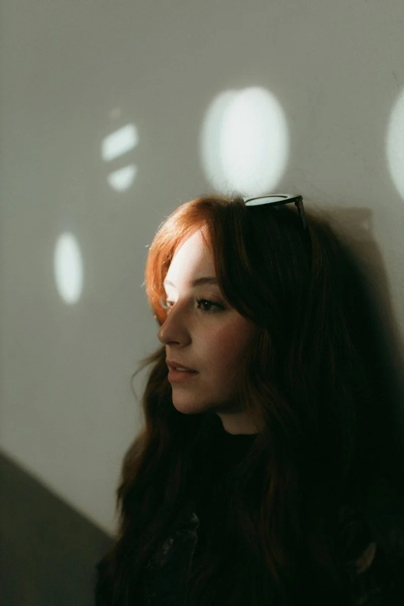 A woman with long, wavy red hair and glasses resting on her head, standing against a wall with light and shadow patterns on her face and hair.