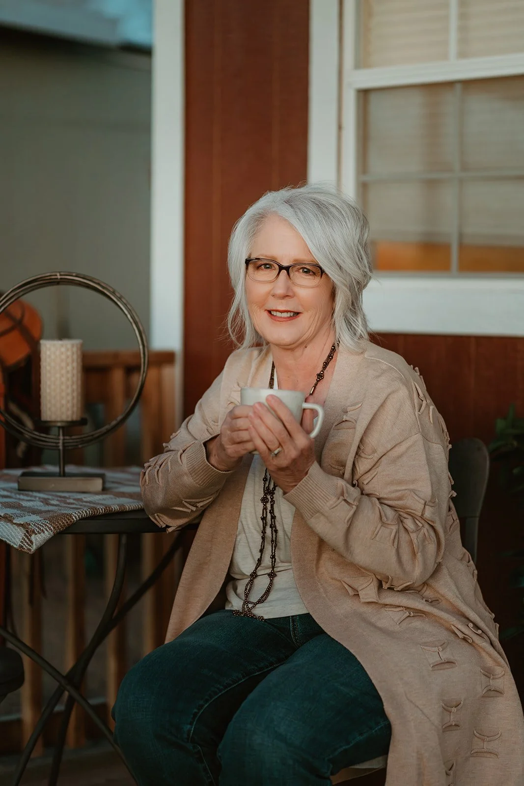 Older woman with gray hair and glasses sitting at a table, holding a mug and smiling.