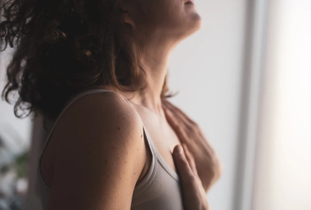Close-up of a woman with curly hair, wearing a tank top, holding her hand on her chest, looking at a bright window.