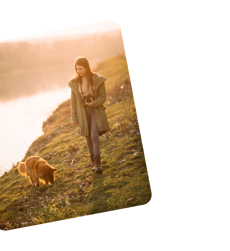 A woman walking along a grassy hillside during sunset, holding a camera, with a small brown dog nearby.