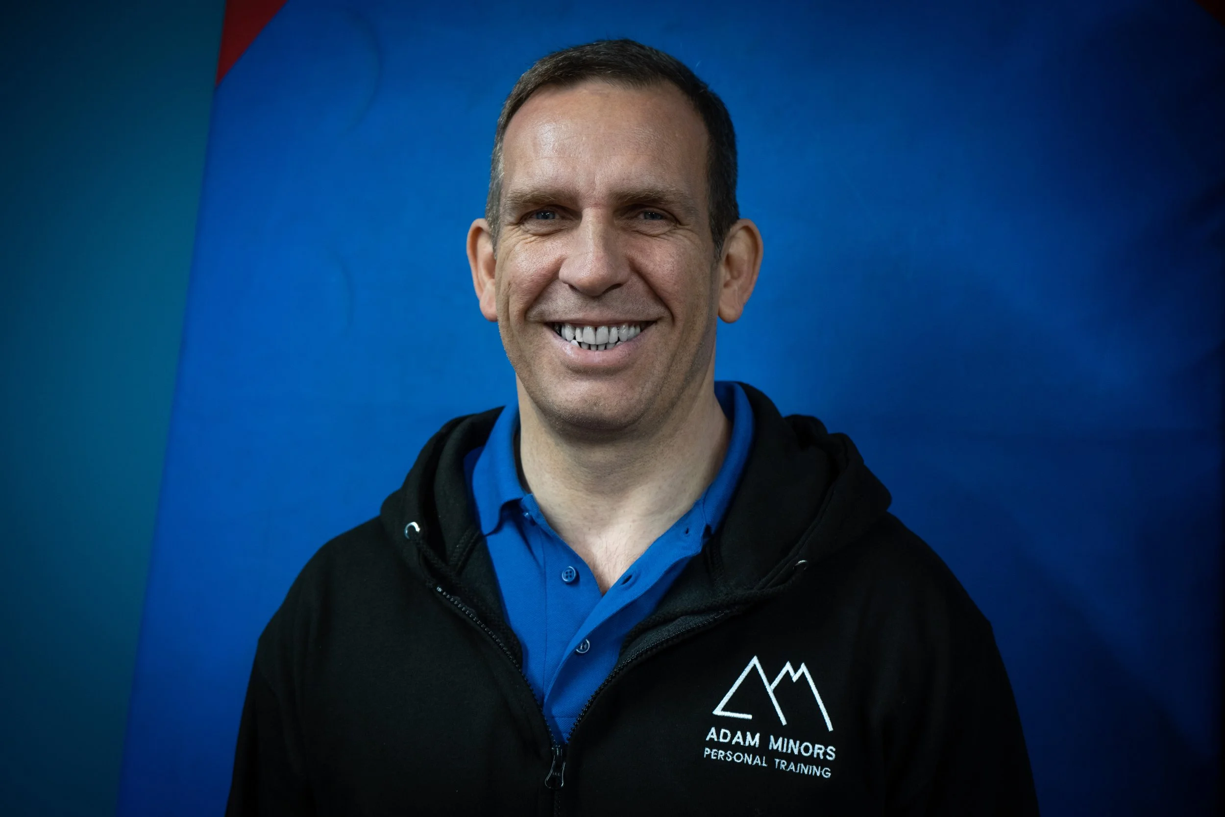 Smiling man wearing a blue polo shirt and a black jacket with a logo and text that reads 'Adam Minors Personal Training' against a blue background.