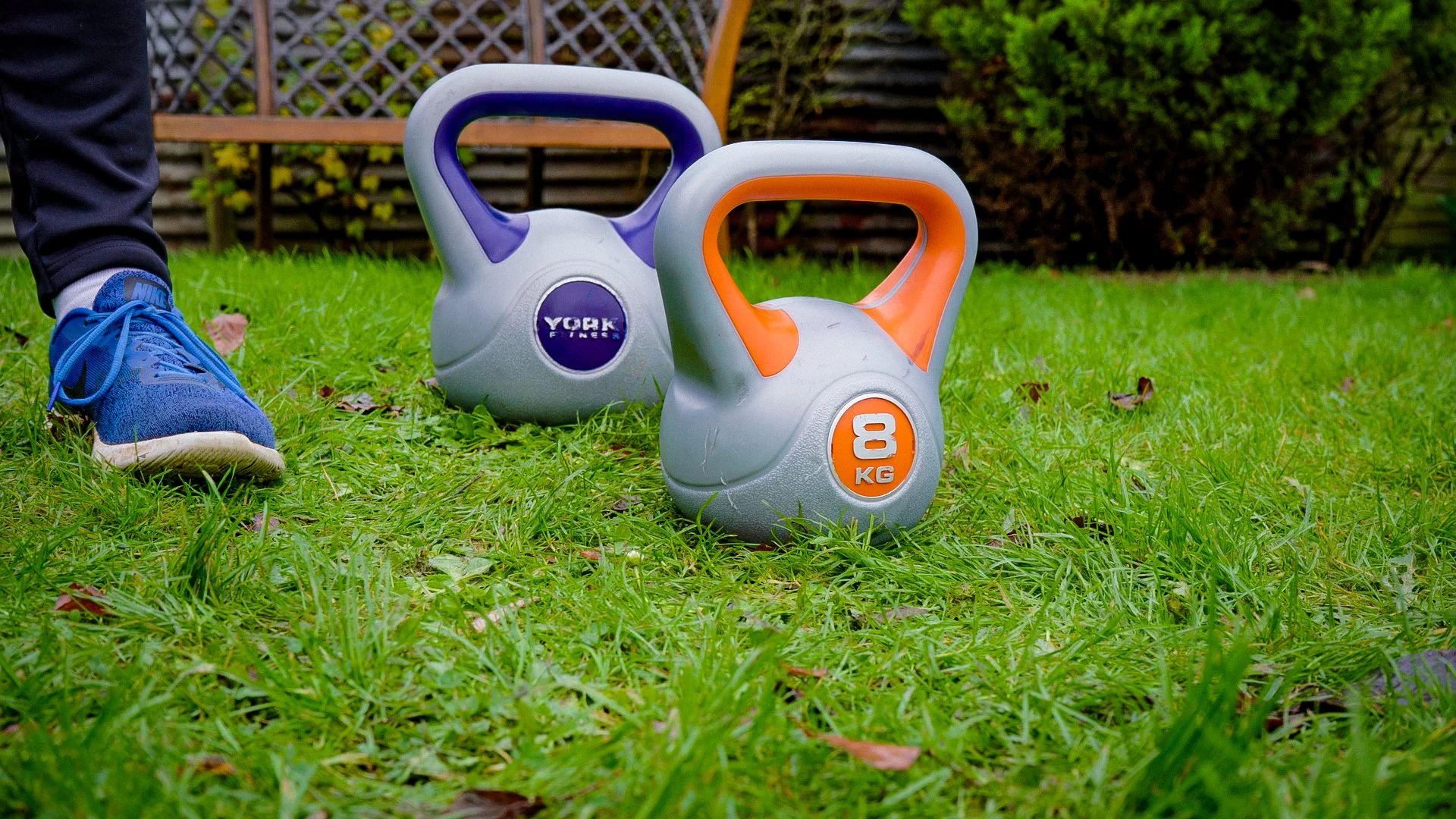 Two kettlebells on a grassy lawn, one with purple accents labeled "York Fitness" and one with orange accents labeled "8 KG." A person wearing blue Nike sneakers and black pants stands nearby.
