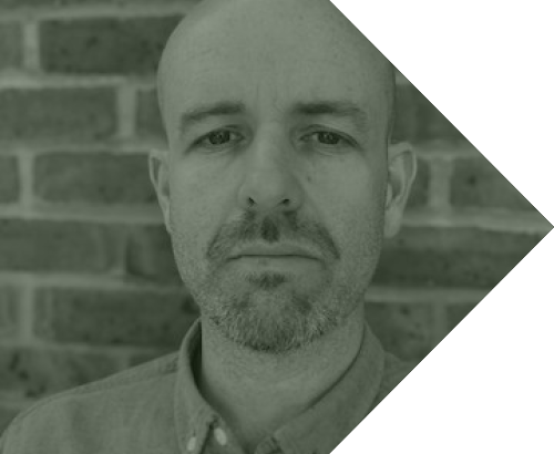 Close-up of a bald man with a beard, wearing a button-up shirt, standing in front of a brick wall.