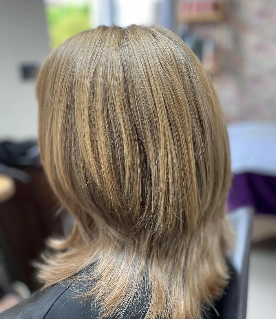Back of person's head with shoulder-length straight blonde hair, indoors with bookshelves and windows in the background.
