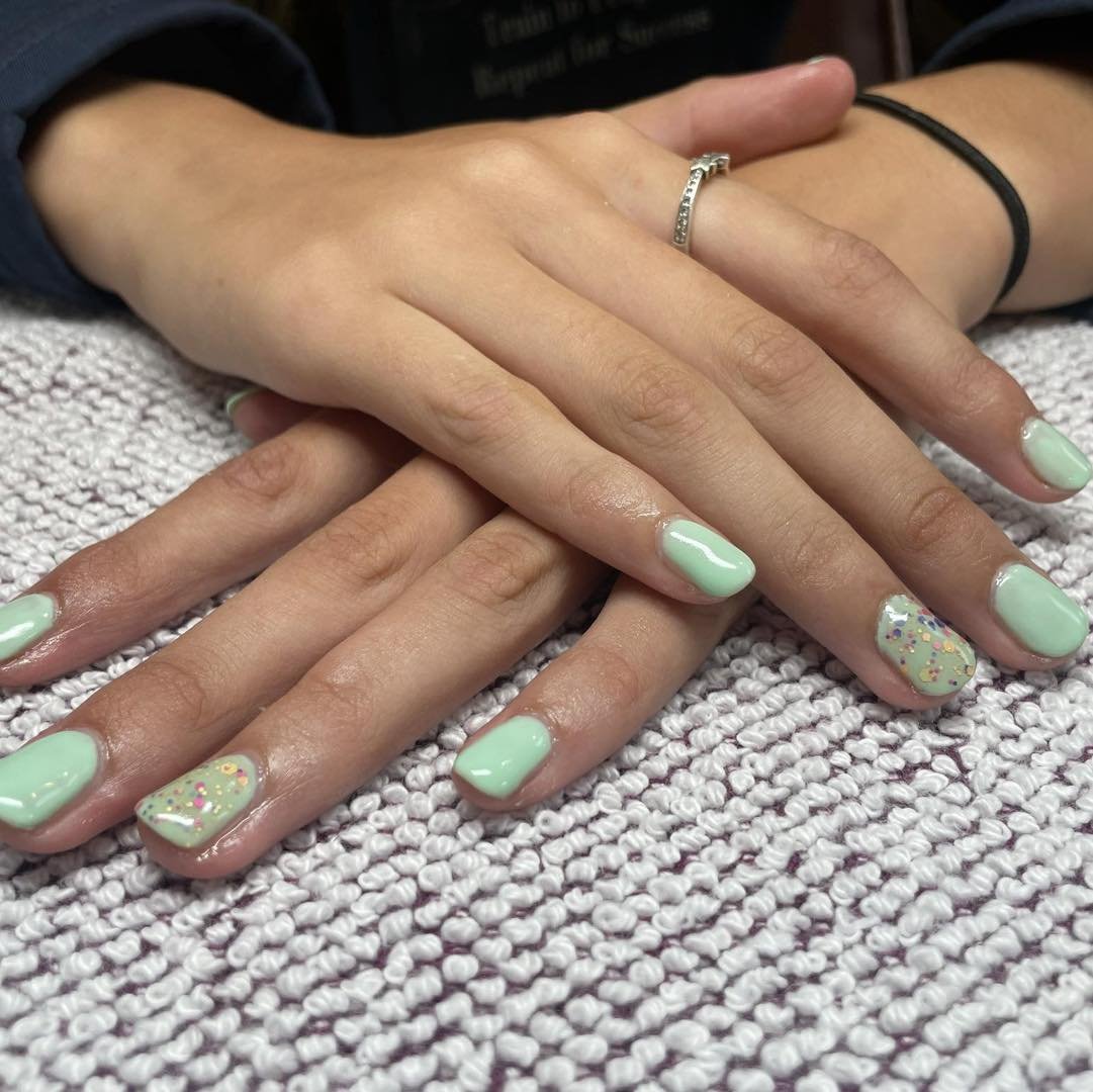 Close-up of manicured hands resting on a textured surface, with mint green nail polish and colorful glitter accent nails.