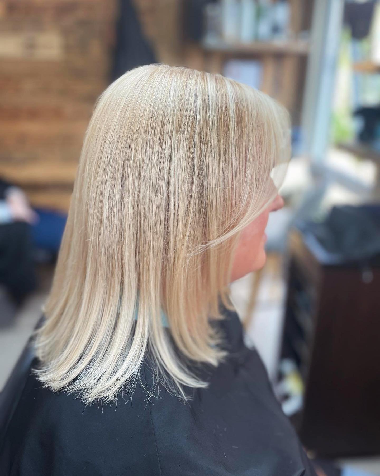 Blonde woman with straight hair in a salon or studio, with a blurred background of wooden shelves and a window.