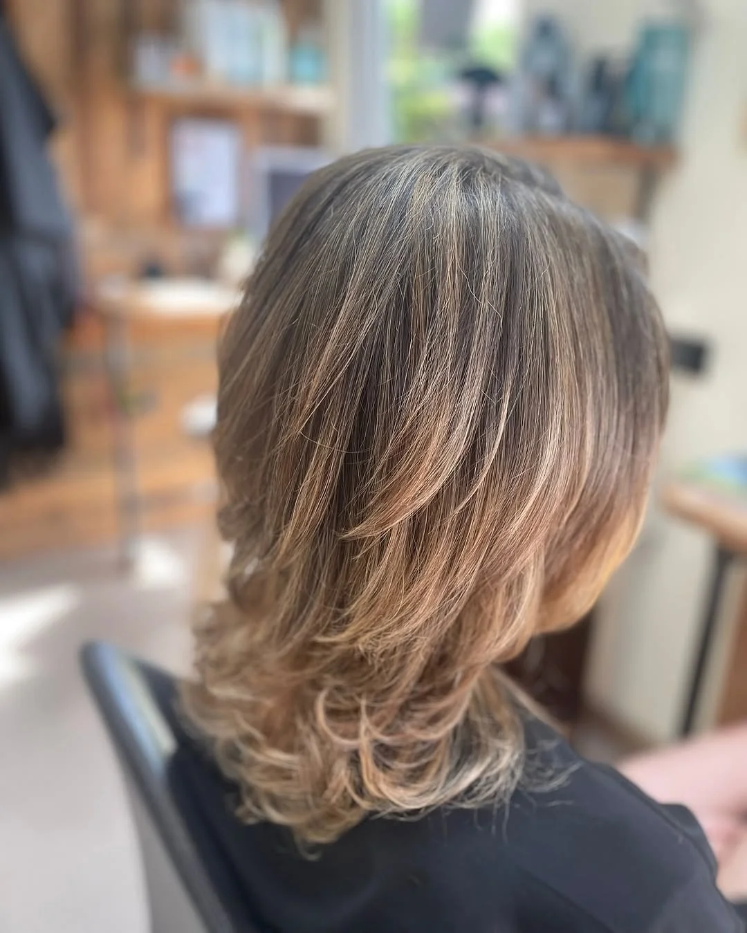 Back view of a woman with light brown hair, styled in loose waves, sitting in a salon chair.