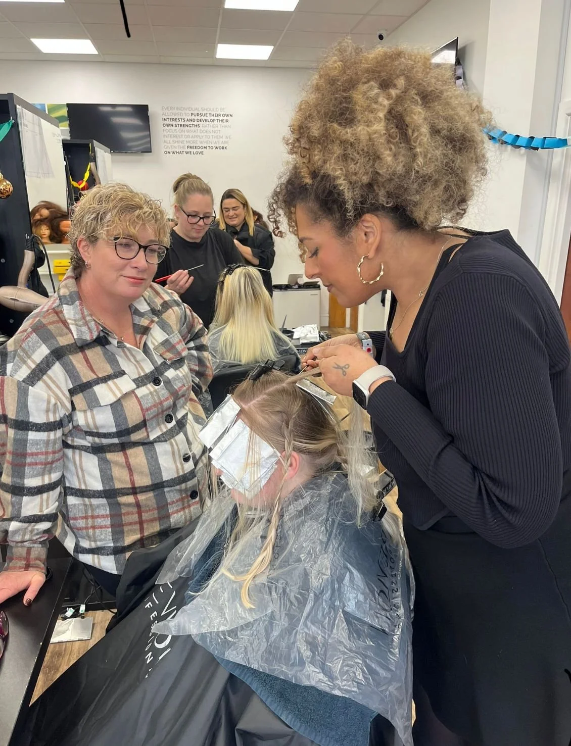 A woman with curly blonde hair getting her hair colored or styled by a hairstylist, with a group of people in the background inside a salon.
