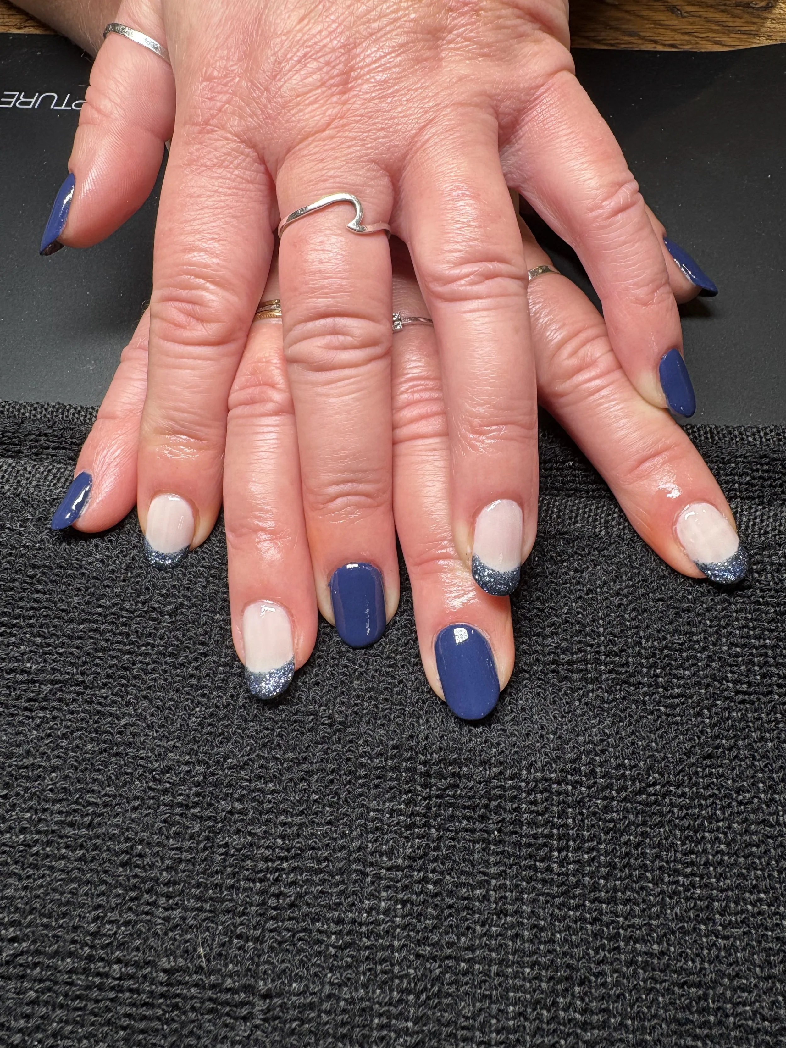 Close-up of hands with manicured nails painted in dark blue and glittery silver tips. One hand shows multiple rings, including a silver wave-shaped ring. The background is black and textured.