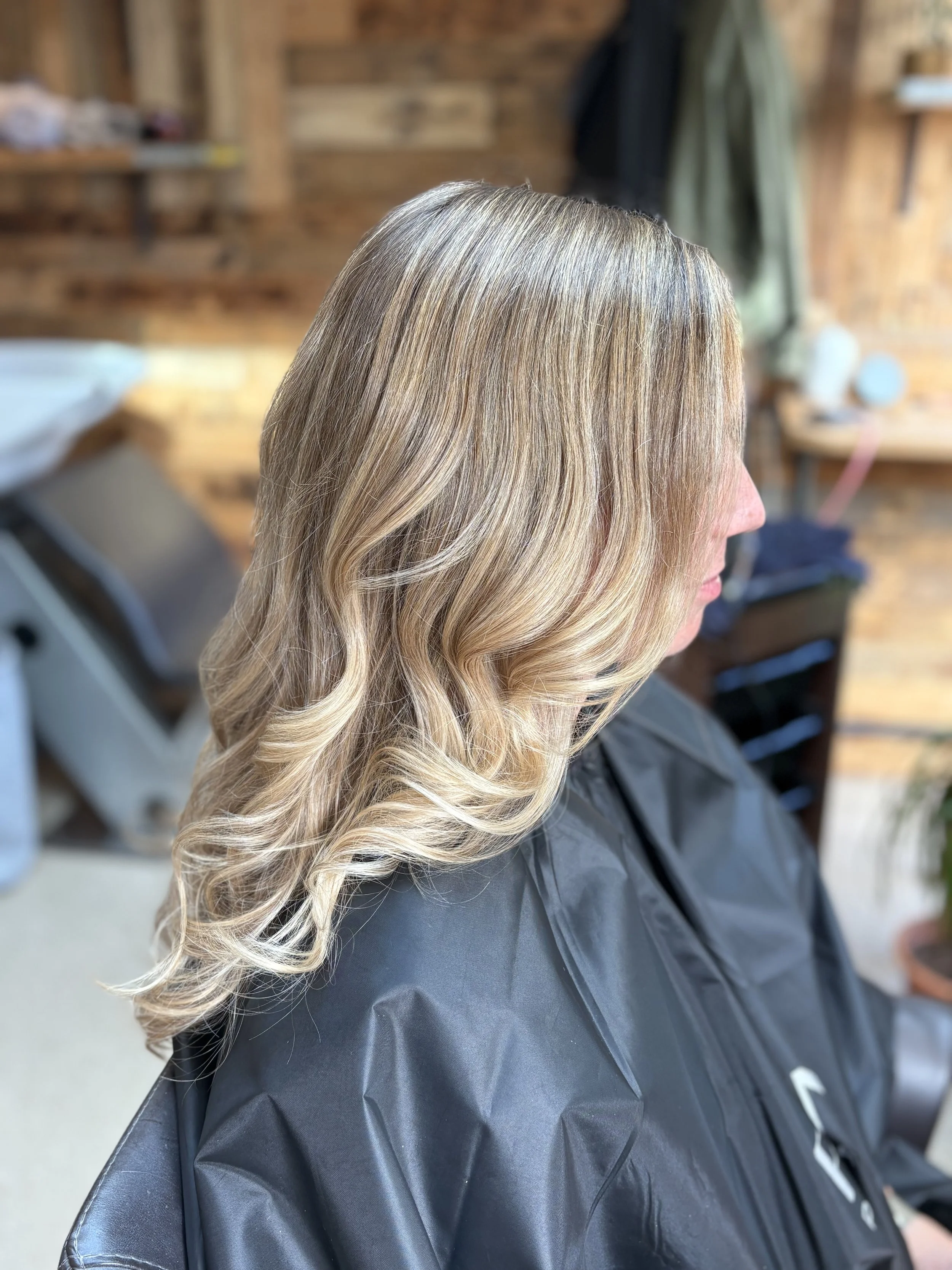 Close-up of a woman's blonde hair with loose curls, sitting in a salon chair with a black apron and a wooden wall background.