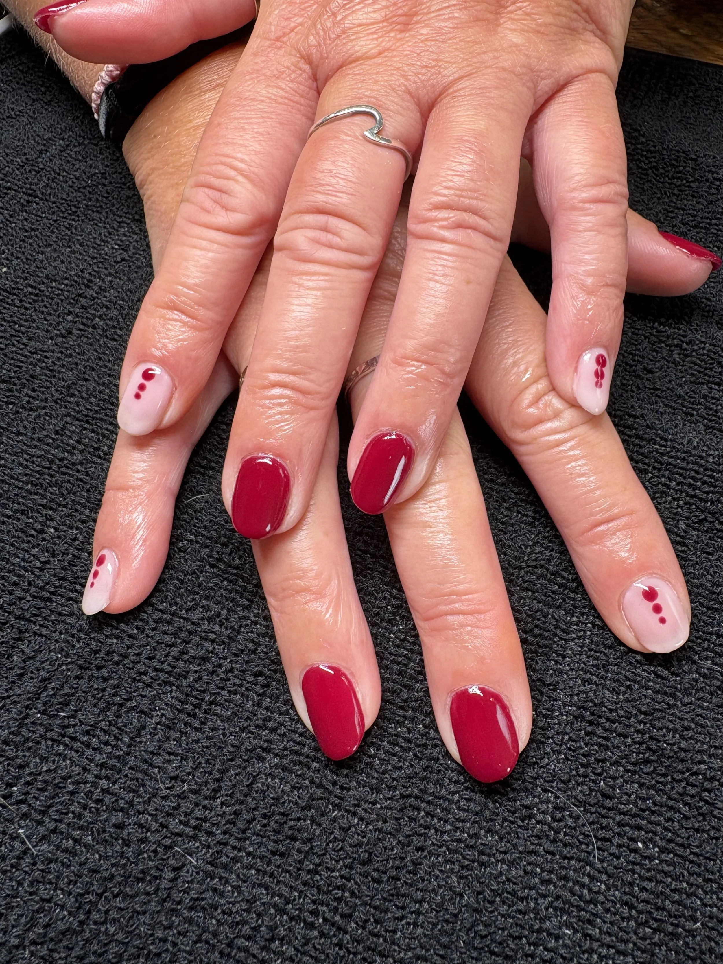 Close-up of a person's hand with red and white nail polish featuring small dot and circle designs, resting on a black textured surface.