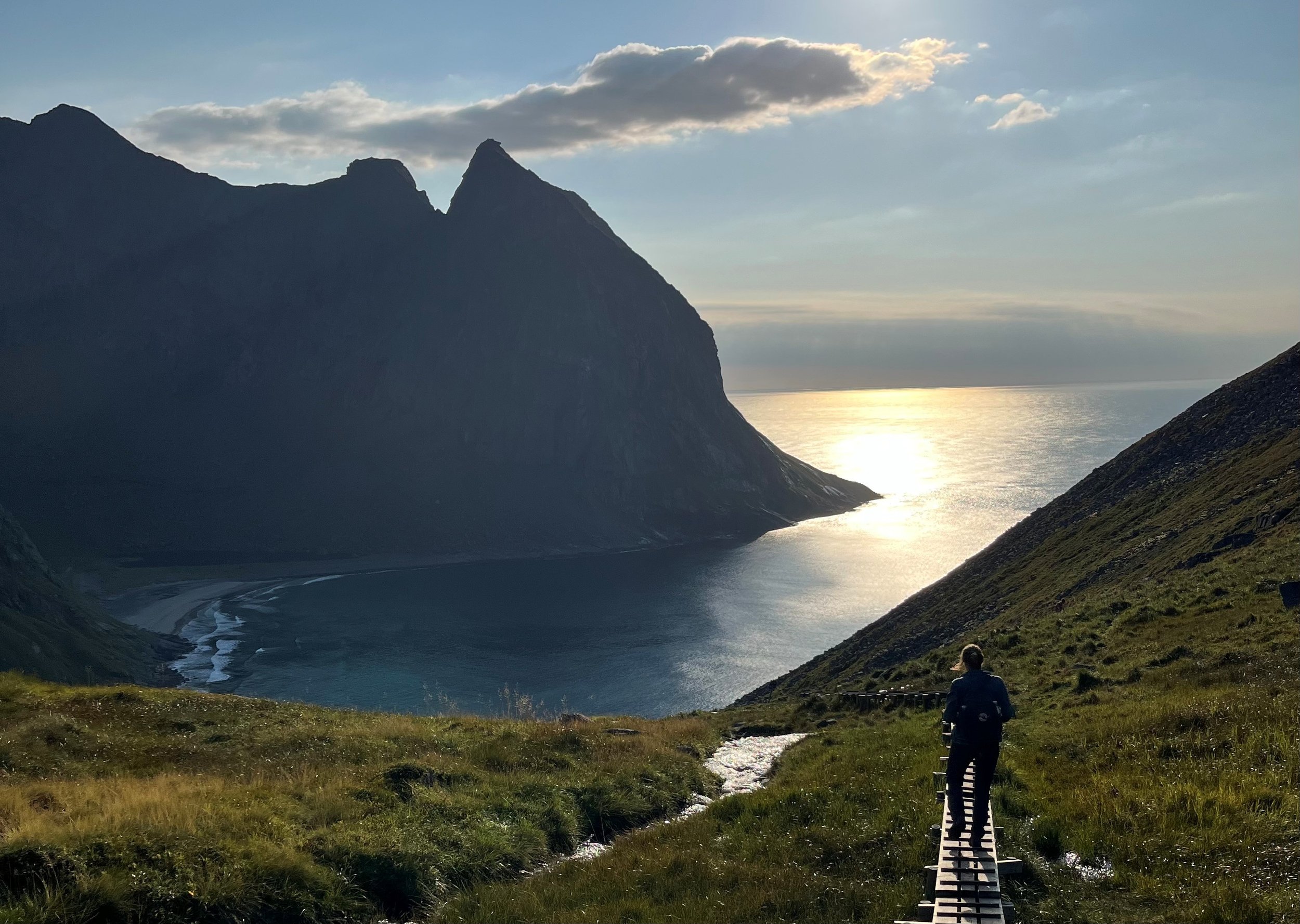 En person går i en fjellandskap med solnedgang eller soloppgang, fjell og grassletter.