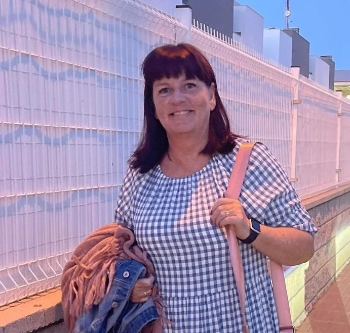 Kvinne smilende foran en hvit gitterveg og lyseblå himmel, iført blå og hvit rutete kjole og holder en jakke og veske