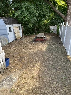 Backyard with a small wooden shed, a picnic table, and a white vinyl fence, surrounded by green trees.
