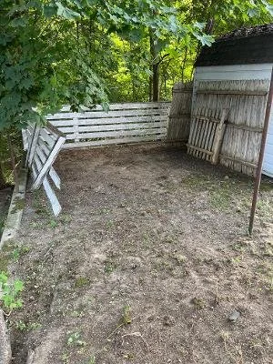A backyard with a dirt ground, white wooden fence, and a small shed with a white and grey exterior, surrounded by green trees.