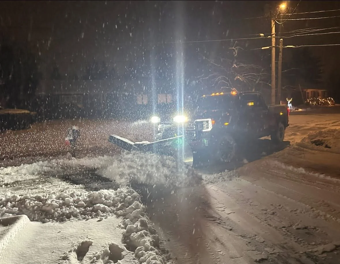A snowplow truck clearing snow from a road at night, with snow falling and being lit by the truck's headlights and a streetlight.