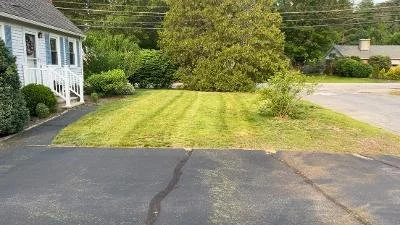 A driveway leading to a small grassy yard with a house on the left and trees in the background.