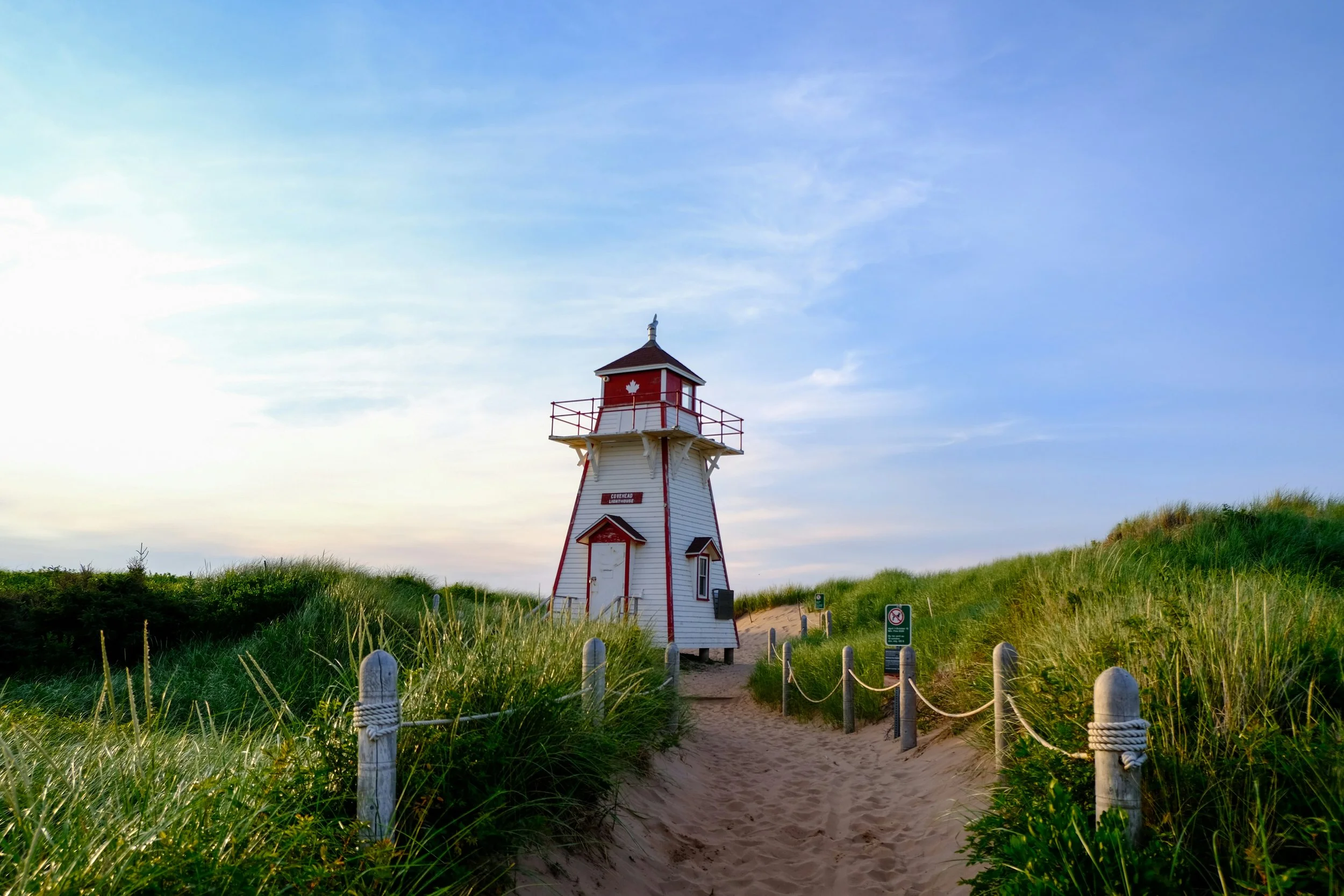 A lighthouse on a sandy path surrounded by green grassy dunes under a blue sky.