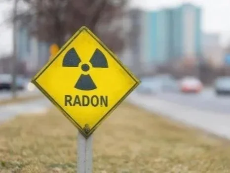 Yellow warning sign with radiation symbol and the word "Radon" below it, located outdoors on a grassy area near a roadway.