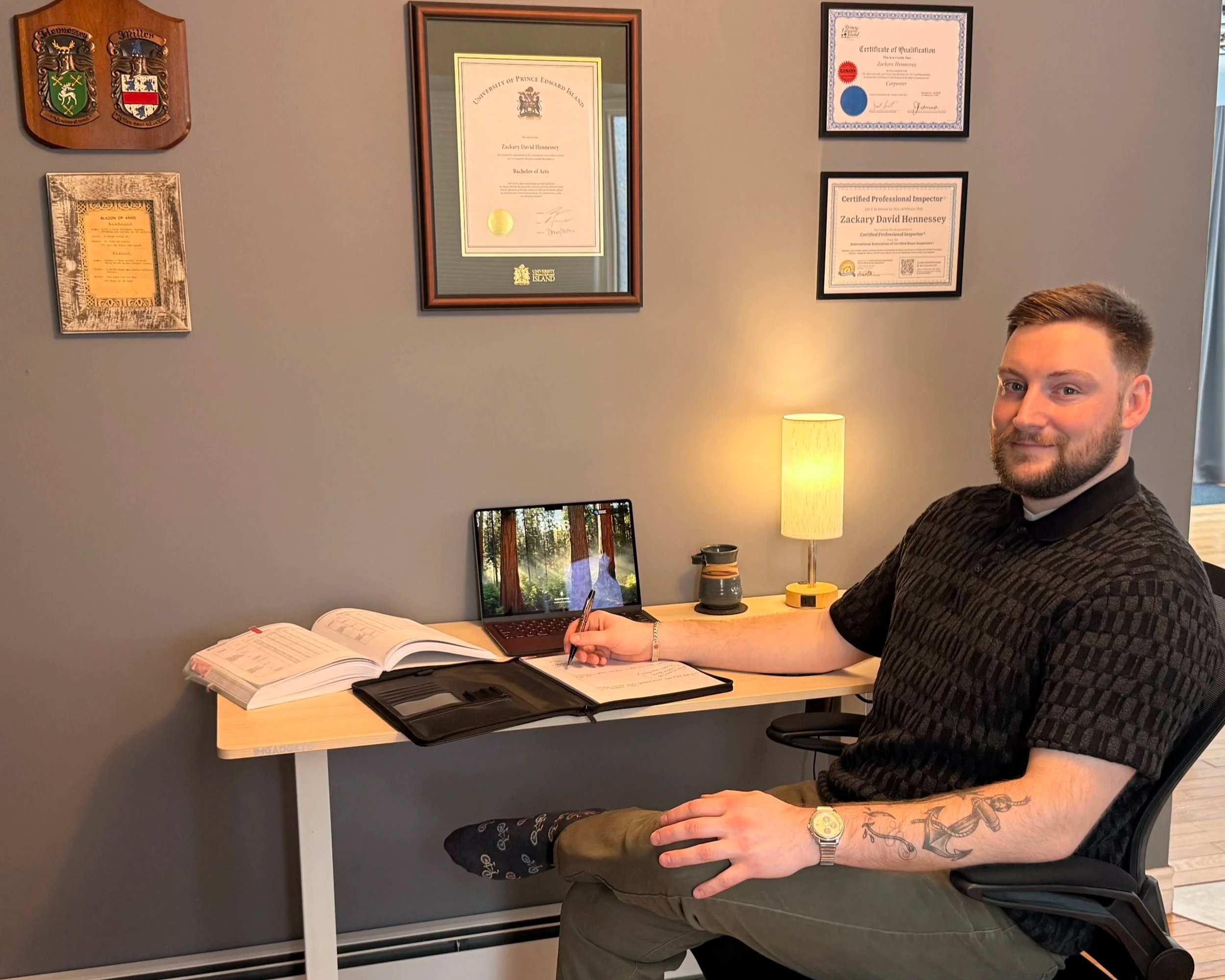 A man sitting at a desk with certificates and awards on the wall behind him, writing in a notebook, with a lamp and a laptop on the desk.