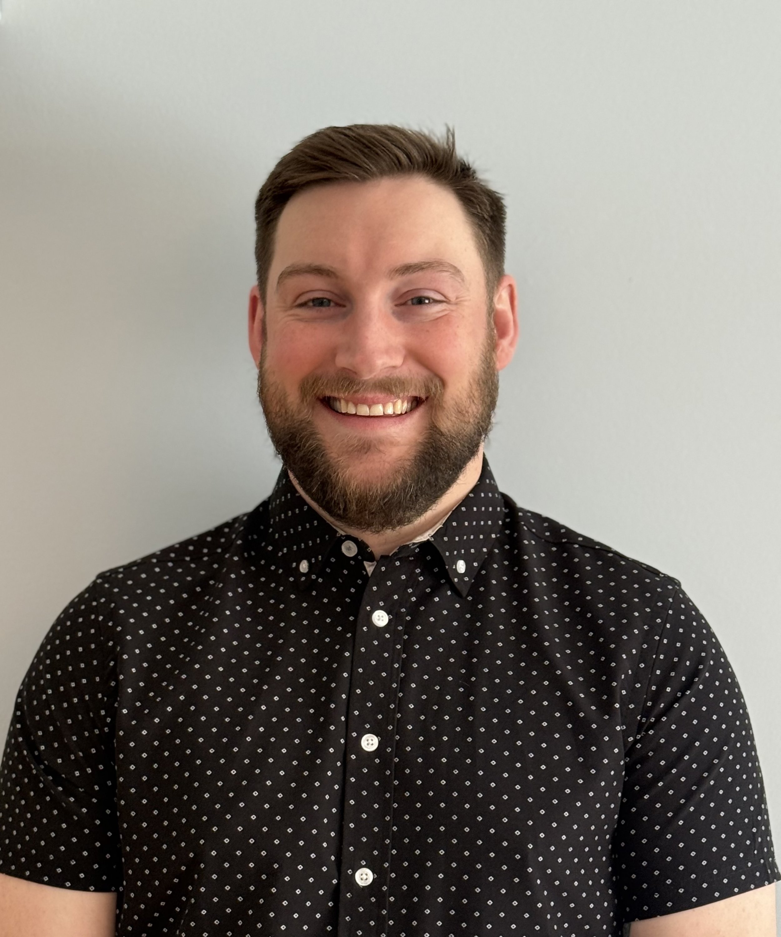 A smiling young man with a beard and short brown hair, wearing a black shirt with white polka dots, standing against a plain light gray background.