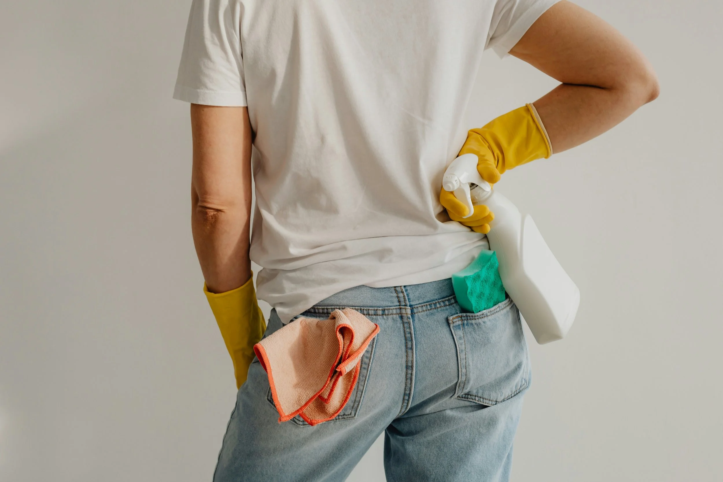 Person wearing a white t-shirt and jeans holding cleaning supplies, including spray bottles, a sponge, and a cleaning cloth, with yellow gloves.