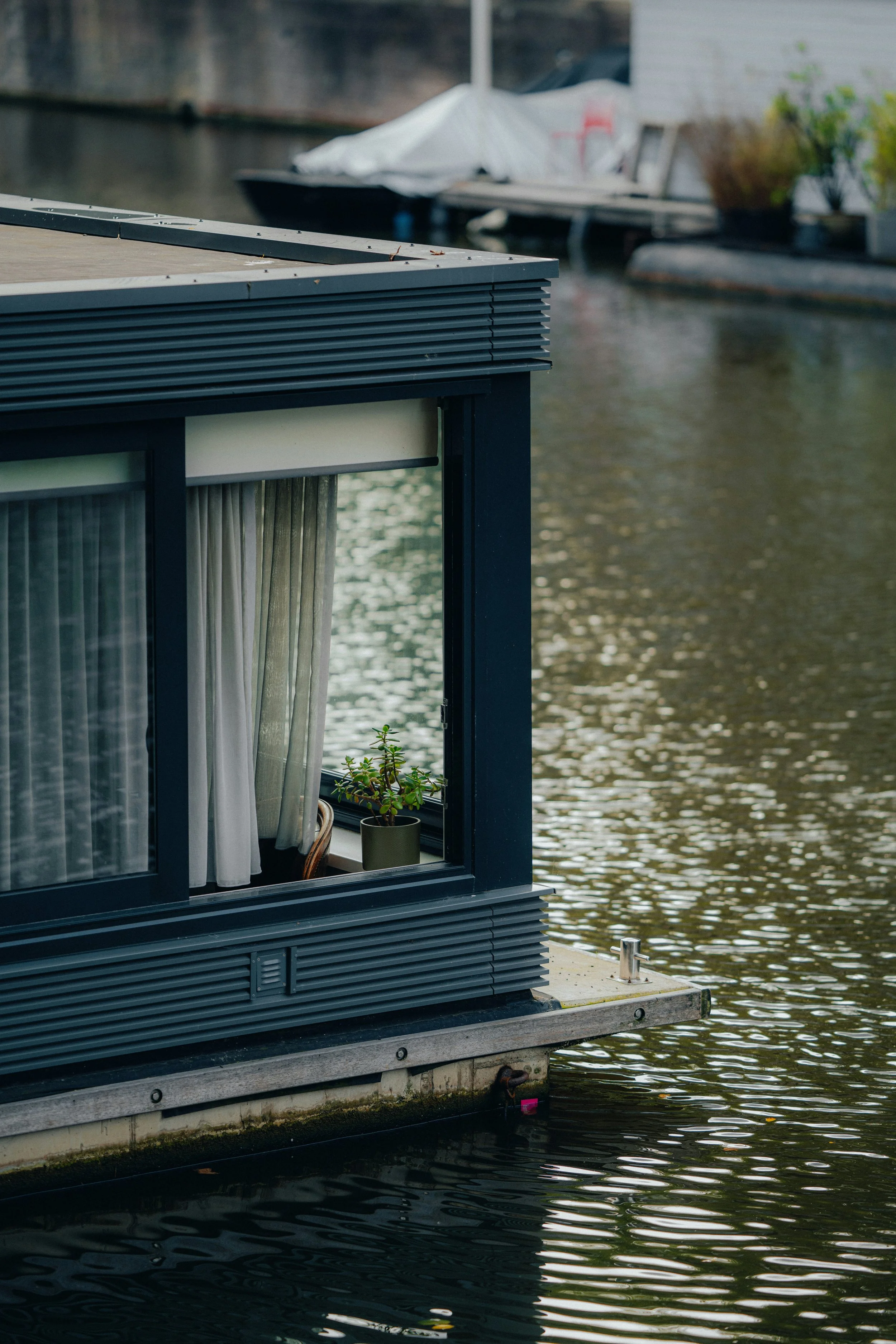 Close-up of a houseboat with large windows, white curtains, a potted plant inside, floating on a river, with another boat docked in the background.