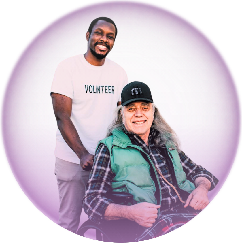 A young man smiling and wearing a white volunteer t-shirt is standing behind an older woman sitting in a wheelchair. The woman is wearing a black baseball cap, a green vest, and a plaid shirt. They are both smiling against a pinkish-purple background.