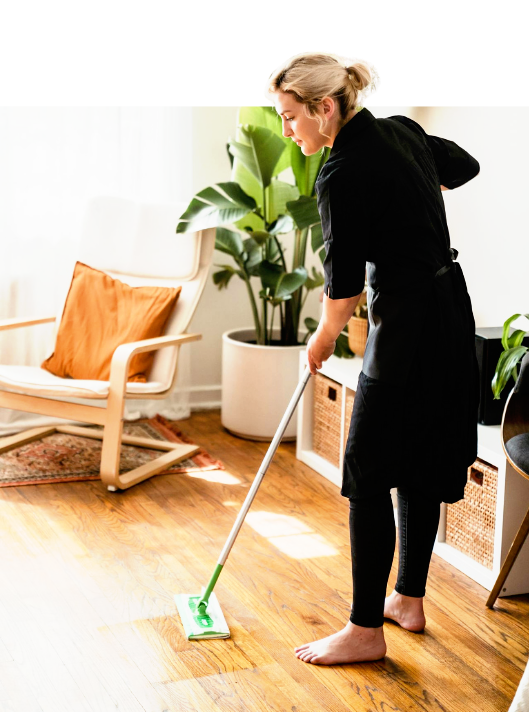 A woman mopping hardwood floor in a living room with plants, a chair with an orange pillow, and natural light.