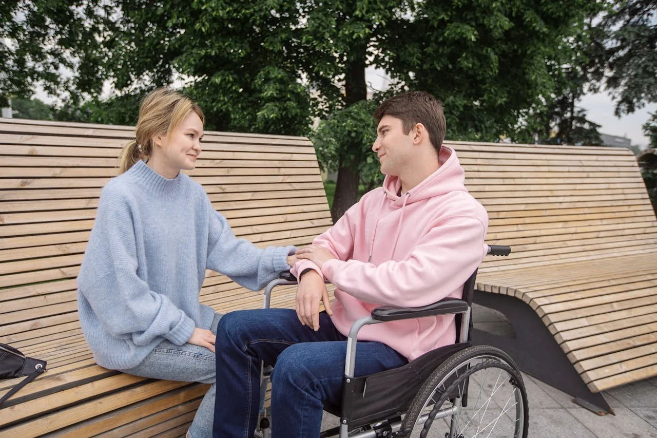 A woman with a light blue sweater sitting on a wooden bench, holding the hand of a man in a pink hoodie who is sitting in a wheelchair. They are outdoors with trees in the background.